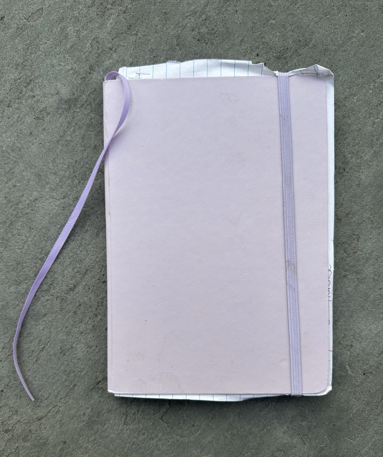 A lavender hardcover notebook on a slate surface, closed with a matching elastic band, its ribbon bookmark trailing off to one side and a few loose ruled pages poking out of the top